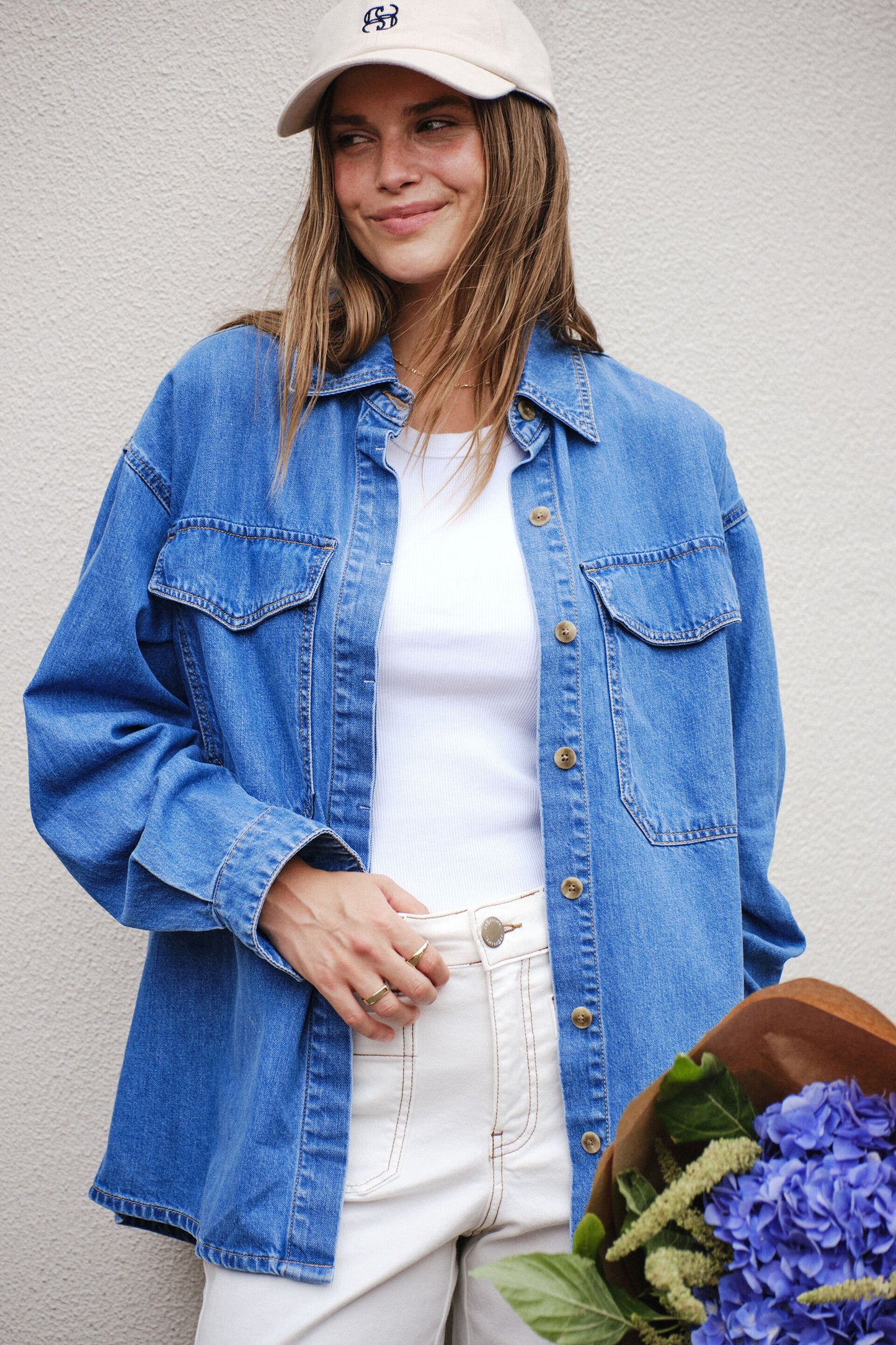 Behind The Trees - Woman wearing a blue denim jacket and white cap, holding flowers against a plain background - Ceres Life - Essential Denim Shirt - Mid Blue Wash