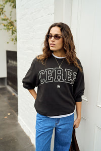 cBehind The Trees - A woman wearing a black crewneck sweatshirt with 'CERES' printed in white collegiate-style letters across the chest, paired with blue jeans, standing against a white brick wall. - Ceres Lifestyle - Isla Oversized Slouchy Crew - Phantom/White Heritage Outline