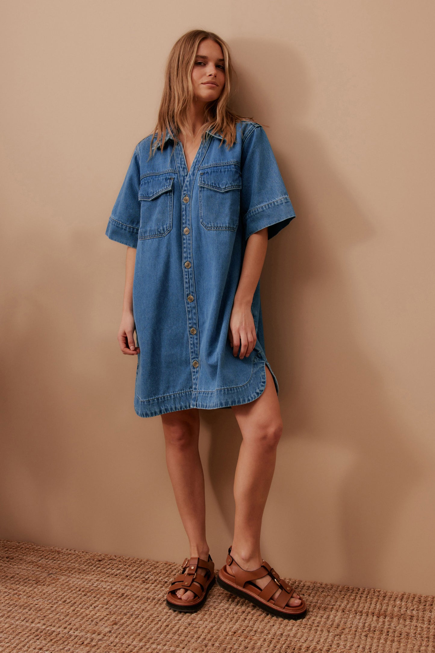 Behind The Trees - Woman wearing a blue denim dress against a beige wall. Ceres Life - Sammy Relaxed Shirt Dress - Fresh Indigo