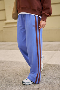 Behind The Trees - Person wearing a blue sweatshirt and pants with a white bag, standing on a wooden floor - Ceres Lifestyle - Gigi Wide Leg Terry Trackpant - Vintage Blue Mixed Side Stripe