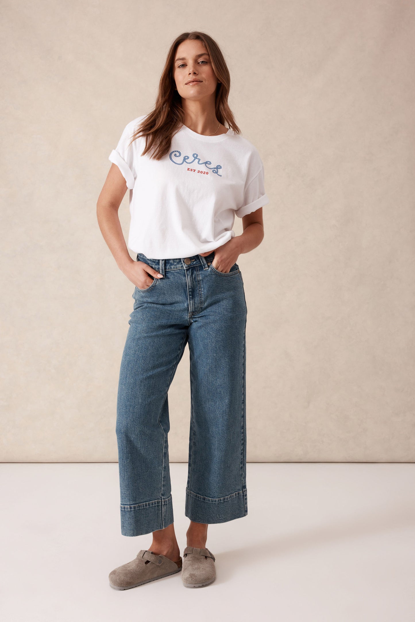 Behind The Trees - Woman wearing a white t-shirt with text and blue jeans against a beige background - Ceres Life - Jude Wide Leg Crop Jean - Mid Indigo