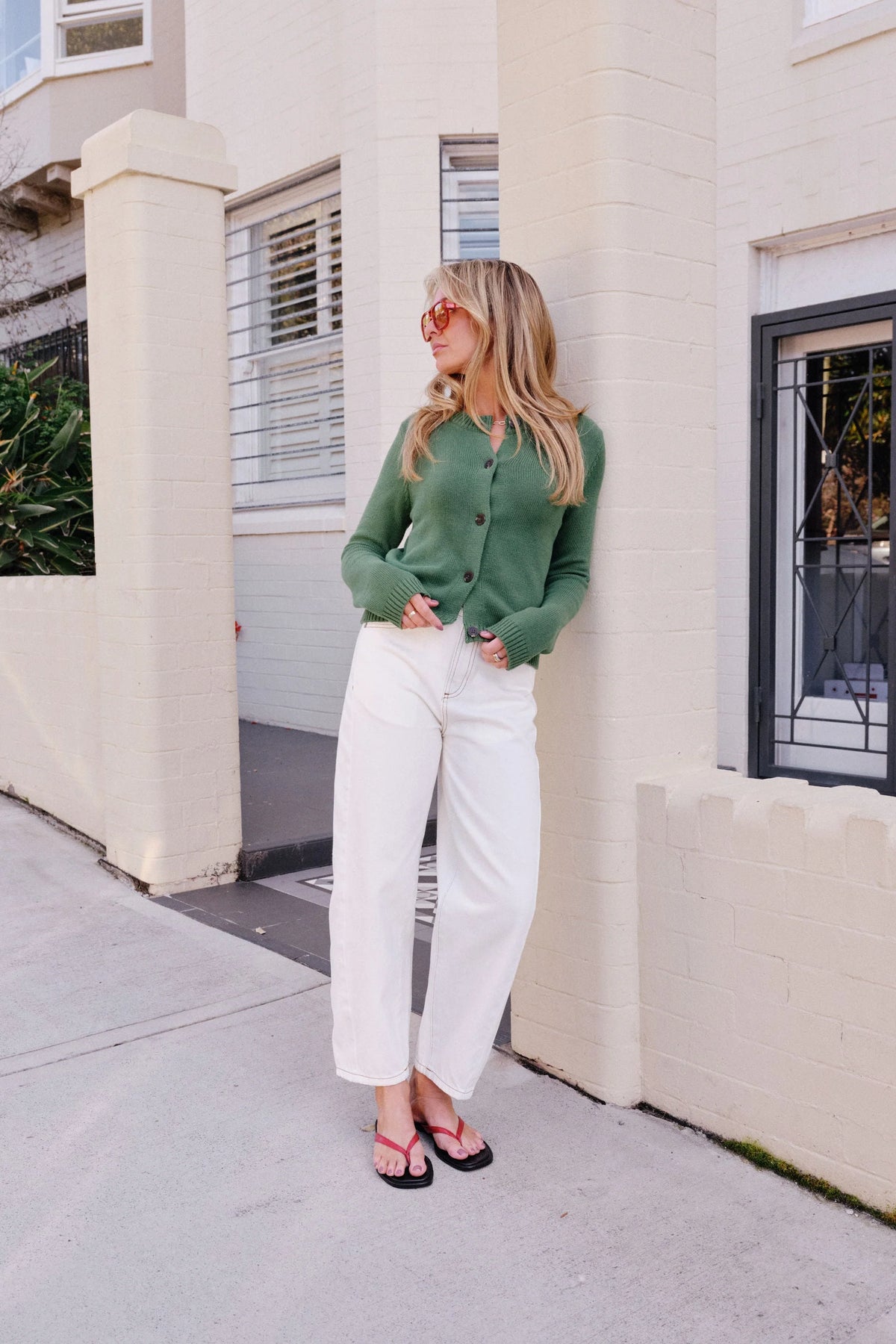 Behind The Trees - A woman wearing a sage green knit cardigan with dark buttons, paired with white pants, standing against a light-colored brick wall. - Ceres Life - Nelly Long Sleeve Knit Cardi - Basil
