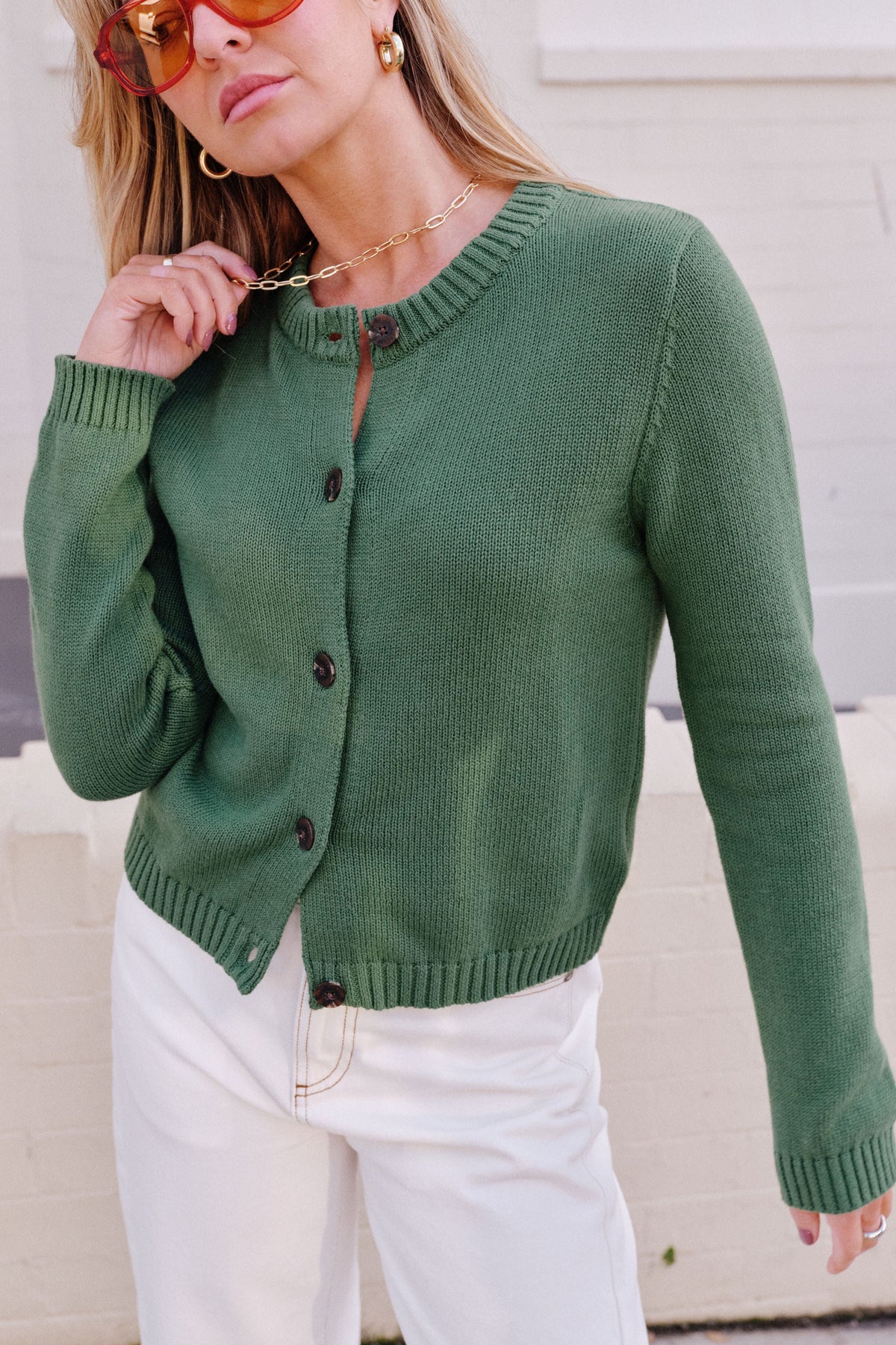 Behind The Trees - A woman wearing a sage green knit cardigan with dark buttons, paired with white pants, standing against a light-colored brick wall. - Ceres Life - Nelly Long Sleeve Knit Cardi - Basil