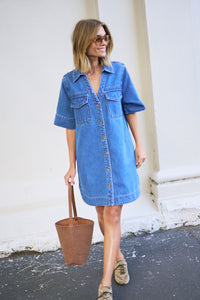 Behind The Trees - Woman wearing a blue denim dress holding a brown bag against a white wall. - Ceres Life - Bonnie Relaxed Shirt Dress -  Fresh Indigo