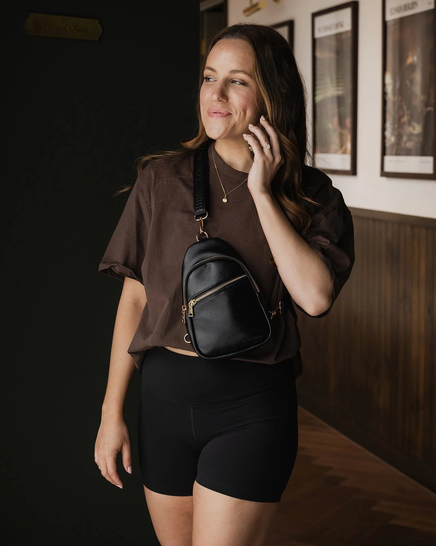Behind The Trees - Woman holding a black backpack indoors with framed pictures on the wall. - Louenhide - Baby Bella Sling Bag - Black
