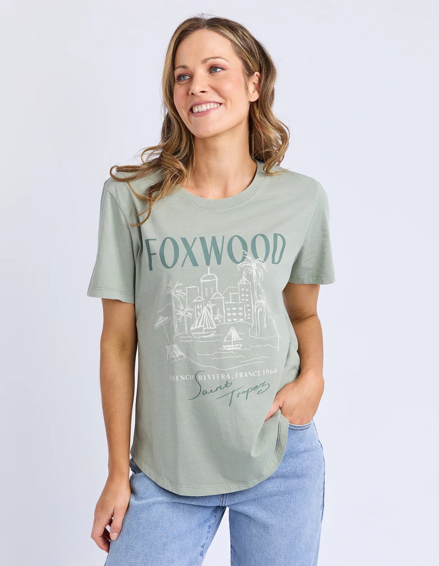 Behind The Trees - Woman wearing a green t-shirt with 'FOXWOOD' printed on it against a light gray background - Foxwood - French Riveria Tee - Iceburg Green