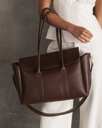 Behind The Trees - Brown leather handbag held by a person wearing a white dress against a gray background - Louenhide - Leighton Tote Bag - Chocolate