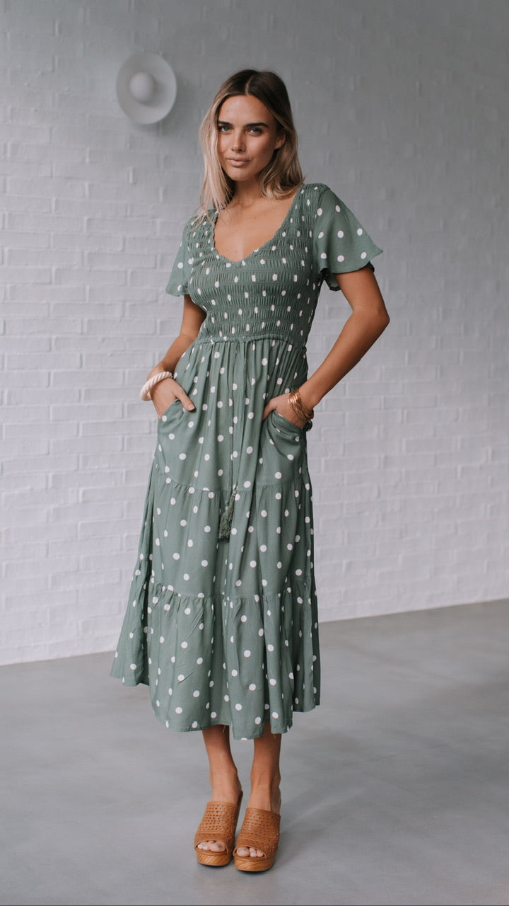Behind The Trees - Woman wearing a green polka dot dress against a gray brick wall. - Grace & Co - Polka Dot Midi Dress - Khaki/White