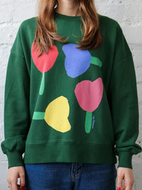 Behind The Trees - Person wearing a green sweatshirt with colorful tulip designs against a white brick wall. - Mosey Me - Forget Me Not Sweater - Forest