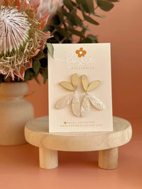 Behind The Trees - Gold earrings on a card with 'Fossil Collective' branding, placed on a wooden stand against an orange background. Foxie Collective - The Love Bug - Gold & Cream Glitter Swirl