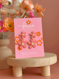 Behind The Trees - Floral earrings on a pink card with 'foxie collective' branding, placed on a wooden stool against a floral background. - Foxie Collective - Double Daisy Dangles - Pastel Raspberry & Gold