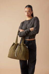 Behind The Trees - A model wearing a dark gray cropped cardigan and black jeans carries an olive green puffer-style drawstring tote bag against a solid beige studio background. - Base Supply - Scrunch Base - Khaki/Cream