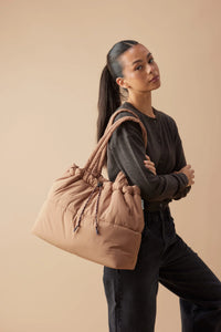 Behind The Trees - A woman with her hair in a ponytail models a large, puffy quilted shoulder bag in a dusty tan color against a solid beige studio background. - Base Supply - Scrunch Base - Hazelnut/Burgundy