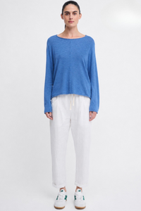 Behind The Trees A model stands against a plain white studio background wearing a heathered blue long-sleeve top, white drawstring pants, and white sneakers with green accents. - Little Lies - Sacha Top - Cornflower Blue