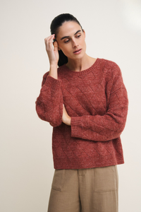 Behind The Trees - A woman wearing a textured, rust-red cable-knit sweater and beige trousers, posing against a plain light beige studio background. - Little Lies - Dylan Jumper - Terracotta