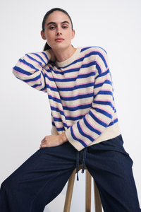 Behind The Trees - A woman seated on a wooden stool wearing a fuzzy long-sleeve sweater with horizontal navy, pink, and cream stripes, paired with dark navy pants, against a plain light gray studio background. - Little Lies - Stripe Otto Knit - Cobalt Stripe