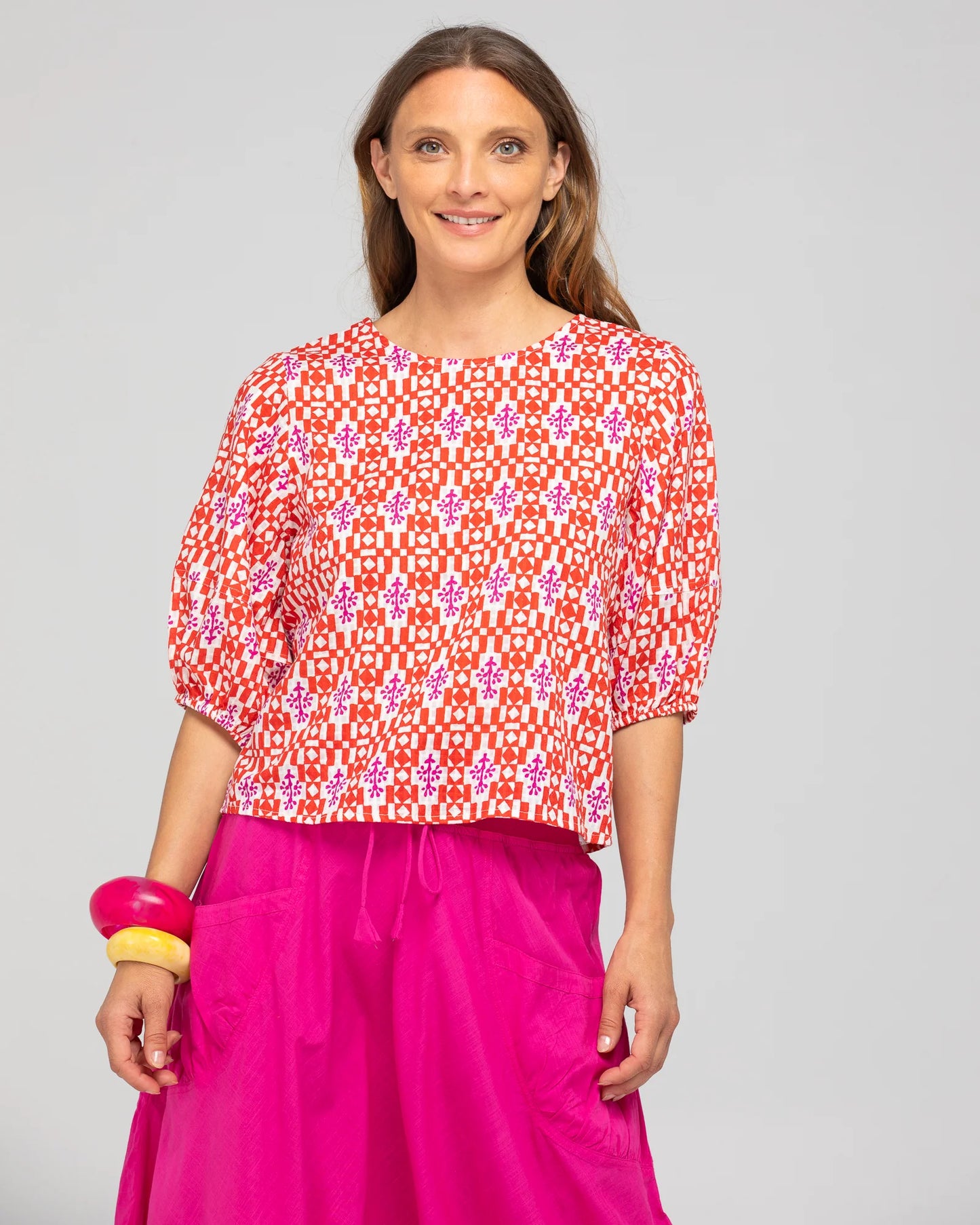 Behind The Trees - Woman wearing a red and white patterned top with pink pants on a gray background - Boom Shankar - Ava Top - Naples Pomodorro