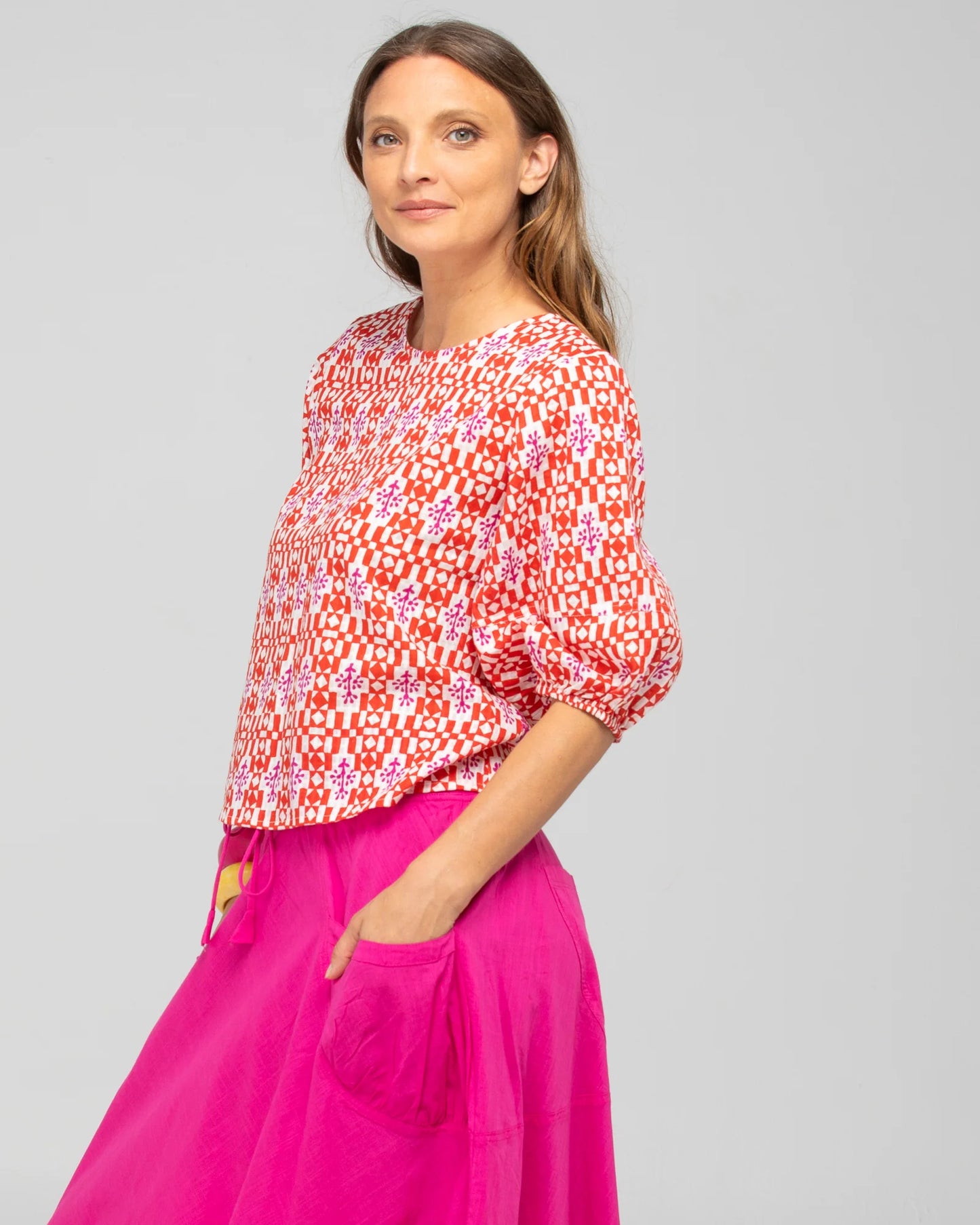 Behind The Trees - Woman wearing a red and white patterned top with pink pants on a gray background - Boom Shankar - Ava Top - Naples Pomodorro