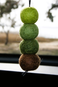 Behind The Trees - A hanging wool felt ornament made of four stacked round balls in shades of green and one brown ball at the bottom, photographed near a window with a blurred outdoor background. - Fresh Ballz - Bush Walkz - Australian Bush Fragrance - Smelly Balls