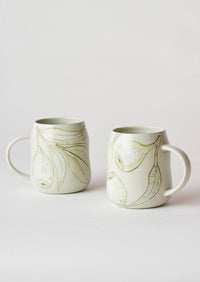Behind The Trees - Two ceramic mugs with a glossy off-white finish, decorated with abstract olive green leaf-like patterns, set against a seamless light gray background. - Angus & Celeste - Everyday Mugs Two Set - Eucalyptus