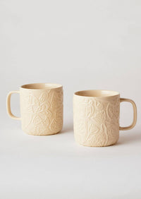 Behind The Trees - Two cream-colored ceramic mugs with embossed floral patterns, shown on a light gray background. - Angus & Celeste - Imaginary Botanical Mugs Two Set - Clay