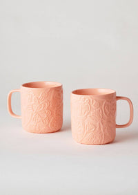 Behind The Trees - Two peach-colored ceramic mugs with embossed floral patterns, displayed on a light gray background. - Angus & Celeste - Imaginary Botanical Mugs Two Set - Coral