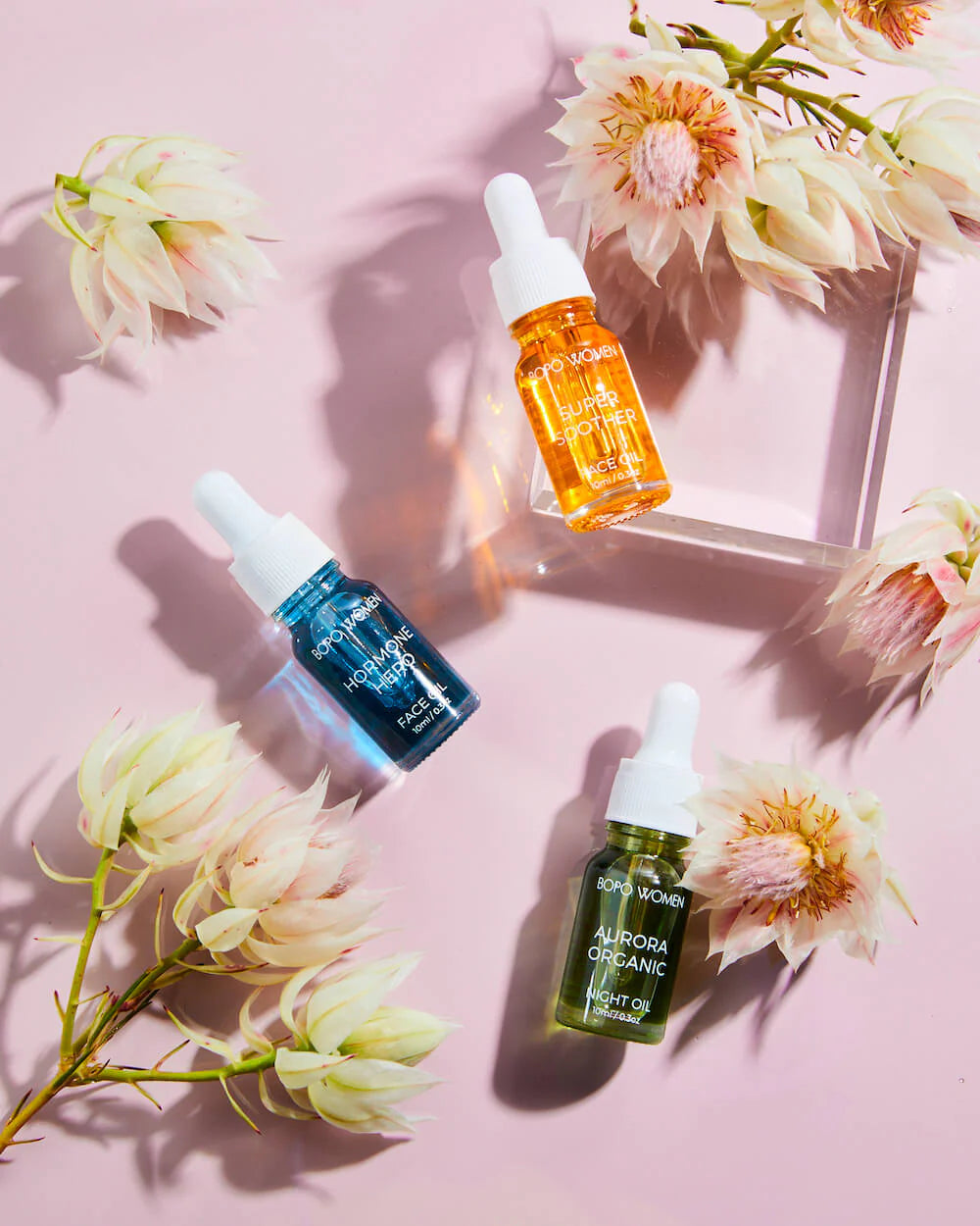 Behind The Trees - Three small glass bottles with droppers containing different face oils, labeled Aurora Organic, Hormone Hero, and Super Soother, displayed in front of a gold foil gift box. - BOPO Women - Face Oil - Mini Sampler Trio