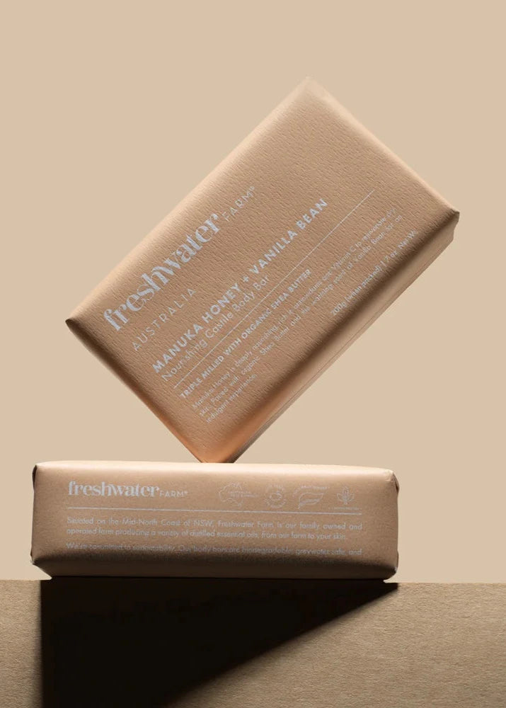 Two Freshwater Farm Manuka Honey + Vanilla Bean Body Bars in elegant, beige-toned recyclable paper packaging. One bar rests flat while the other leans diagonally against it, set against a neutral-toned background. The packaging highlights key product details, including its nourishing formula with organic shea butter, triple-milled craftsmanship, and commitment to sustainability with biodegradable and greywater-safe ingredients. Available for purchase from Behind The Trees.