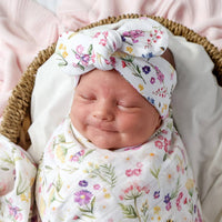 Behind The Trees-  A sleeping newborn baby swaddled in a white muslin blanket with a colorful wildflower print, wearing a matching bow headband, resting inside a woven wicker basket on a soft pink blanket. Snuggle Hunny - Organic Topknot - Gracie