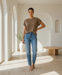 Behind The Trees - Woman wearing a brown t-shirt and blue jeans standing in a bright, minimalistic room. - Little Lies - Rhodes Tee - Mocha