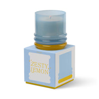 Behind The Trees - Candle in a blue container with a box labeled 'Zesty Lemon' on a white background - Designworks - Stacked 7oz Glass w/ Yellow Lid and Blue Base - Zesty Lemon