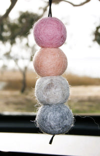 Behind The Trees - A hanging felt car ornament made of four stacked wool balls in pink, peach, white, and blue-gray, suspended by a black string in front of a blurred car window. - Fresh Ballz - Shepard's Delight - Musk Sticks Fragrance - Smelly Balls