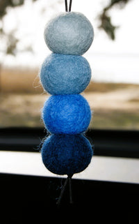 Behind The Trees - A handmade felt wool hanging ornament made of four stacked oval felt balls in a vertical gradient from light blue to dark navy blue, suspended by a black string in front of a blurred window. - Fresh Ballz - Winter Wavez - Coconut & Lemongrass Fragrance - Smelly Balls
