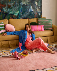 Behind The Trees - Woman in a pink dress and blue robe sitting on a yellow couch with colorful pillows and a painting in the background. - Kip & Co - Adult Cosy Robe - Pie In The Sky