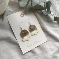 Behind The Trees - Pair of gold and white earrings on a card with 'Foxie Collective' branding, placed on a textured surface. - Foxie Collective - Marlie Earrings - Gold Glitter & Cream