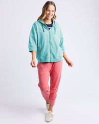 Behind The Trees - A woman modeling a light aqua zip-up hoodie and coral pink jogger pants against a plain light gray studio background. - Elm - Beachy Zip Hoodie - Tonic Blue