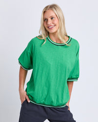 Behind The Trees - A smiling blonde woman wearing a bright green short-sleeve t-shirt with white and green striped trim on the collar and sleeve cuffs, paired with dark pants, against a light gray studio background. - Elm - Faye Short Sleeve Sweat - Green/Navy & White