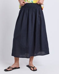 Behind The Trees - A person wearing a dark navy blue linen midi skirt with a smocked elastic waistband, paired with black flip-flops and a colorful floral top, shown against a light gray studio background. - Elm - Flair Shirred Skirt - Navy 