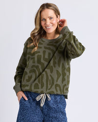Behind The Trees -Woman wearing a green camouflage sweater and blue leopard print pants on a white background -Elm - Gala Crew - Clover Yardage Print
