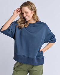 Behind The Trees - Woman wearing a blue sweatshirt and green pants on a light gray background - Elm - Ines Sweat - Navy