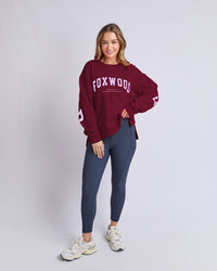 Behind The Trees - Woman wearing a maroon 'FOXWOOD' sweatshirt and blue leggings on a light gray background -Foxwood - Interval Crew - Port with Lilac
