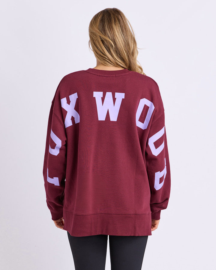 Behind The Trees - Woman wearing a maroon 'FOXWOOD' sweatshirt and blue leggings on a light gray background -Foxwood - Interval Crew - Port with Lilac