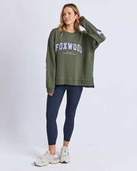 Behind The Trees - Woman wearing a green sweatshirt with 'FOXWOOD' branding and dark blue leggings on a white background - Foxwood - Interval Crew - Khaki with Lilac