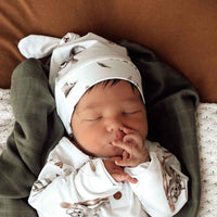 Behind The Trees - Newborn baby wearing a white hat and outfit with animal patterns, lying on a brown blanket. - Snuggle Hunny - Koala Organic Knotted Beanie