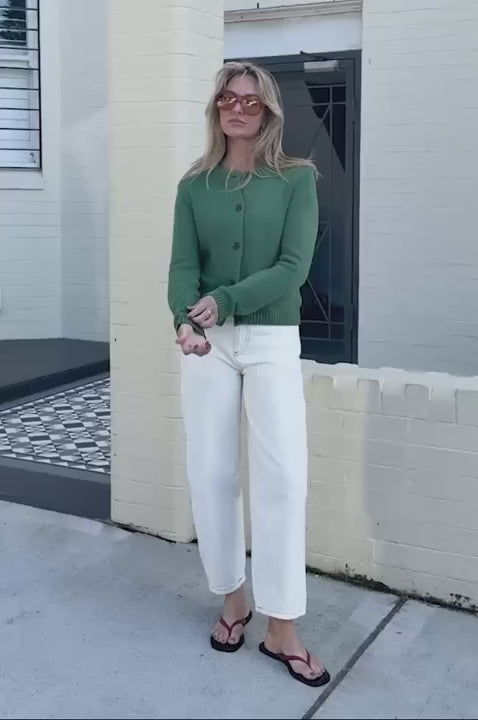Behind The Trees - A woman wearing a sage green knit cardigan with dark buttons, paired with white pants, standing against a light-colored brick wall. - Ceres Life - Nelly Long Sleeve Knit Cardi - Basil
