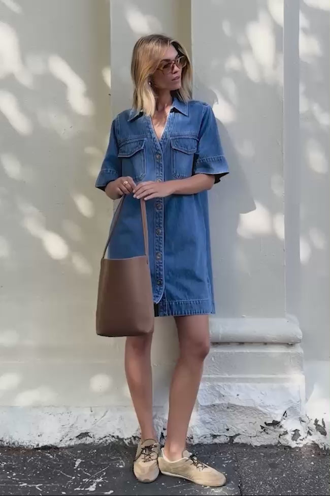 Behind The Trees - Woman wearing a blue denim dress holding a brown bag against a white wall. - Ceres Life - Bonnie Relaxed Shirt Dress -  Fresh Indigo