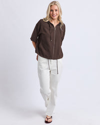 Behind The Trees - A woman modeling a dark brown short-sleeve zip-up hoodie with white drawstring pants and black sandals, standing against a light gray studio background. - Elm - Saltair Lightweight Jacket - Chocolate
