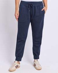 Behind The Trees - A person wearing navy jogger sweatpants with a drawstring waist and elastic cuffs, paired with beige sneakers, shown against a light gray studio background. - Foxwood - Sami Trackpant - Dark Sapphire