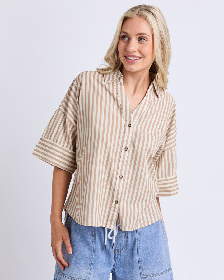 Behind The Trees - A woman wearing a beige and white vertically striped short-sleeve button-up shirt with rolled cuffs, paired with light blue denim shorts, against a light gray studio background. - Elm - Striped Holiday Shirt - Natural